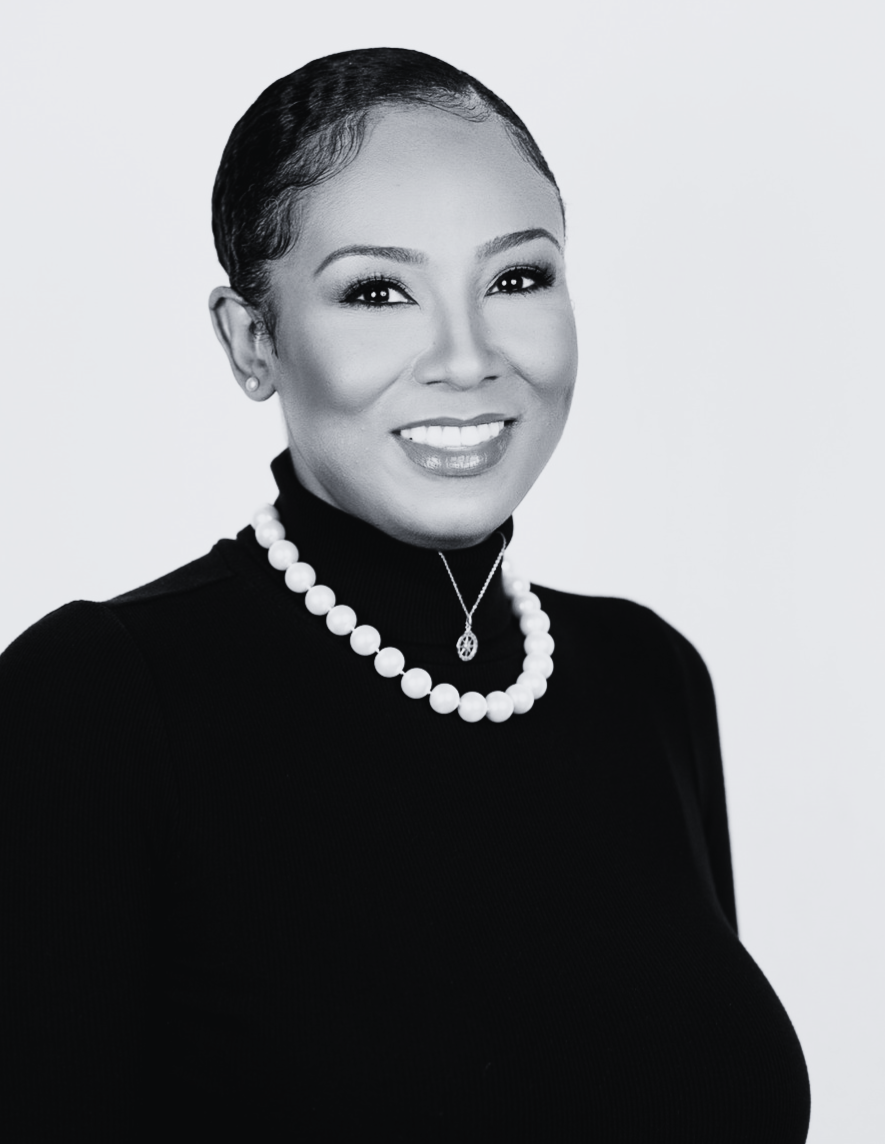 Black-and-white image of Lucky Tigereye wearing a black turtleneck, pearl and compass necklaces, sitting slightly tilted to the side, smiling.